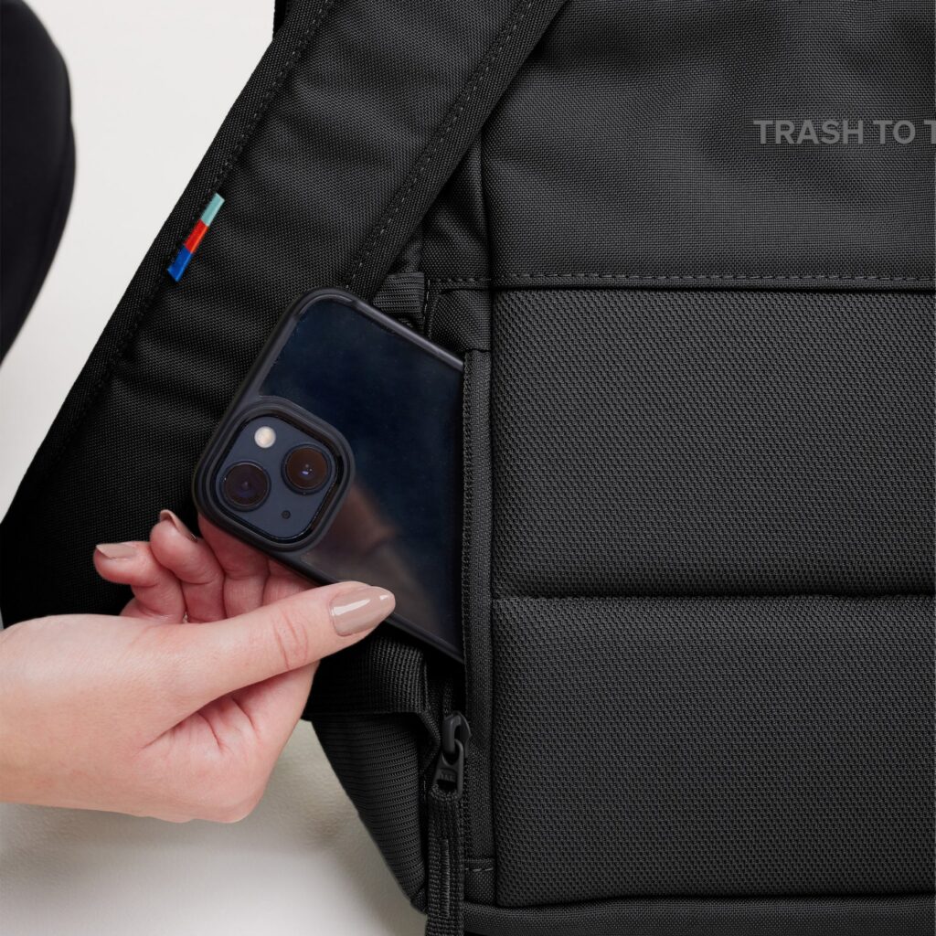 DAYPACK 2.0 Rucksack in black (Schwarz), gehalten von einer Frau, die ihr Telefon in eine Tasche steckt.